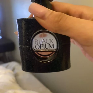 Black Opium Eau de Parfum från Yves Saint Laurent - Säljer en ikonisk Black Opium parfym från Yves Saint Laurent. Flaskan är svart med glittrig yta och rund transparent detalj i mitten där parfymen syns. Märkets logga syns tydligt på flaskhalsen. Perfekt för dig som gillar djupa och sensuella dofter.