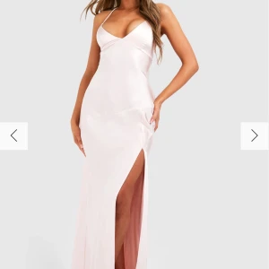 Ljusrosa långklänning med slits och öppen rygg - Elegant ljusrosa långklänning i satin med smala axelband, djup slits framtill och öppen rygg med snörningsdetalj. Klänningen har en figurnära passform och v-ringad urringning, perfekt för speciella tillfällen.