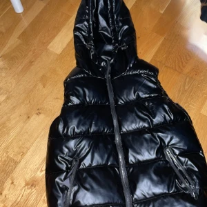 Svart dunväst med huva - Säljer en svart dunväst med huva och dragkedja framtill. Västen har två fickor med dragkedja och är quiltad för extra värme. Perfekt för lager-på-lager under kyliga dagar.