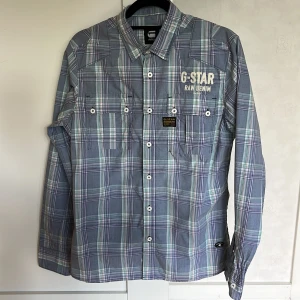 Vintage skjorta från G-Star RAW - Säljer en snygg rutig skjorta från G-Star RAW i blå och vita toner. Skjortan har långa ärmar, knappar framtill och två bröstfickor. Perfekt för dig som gillar streetwear och vill ha något stilrent men ändå avslappnat.