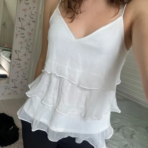 Vitt volanglinne från Vero Moda - Säljer ett vitt linne med tunna axelband och flera lager volanger. Toppen har v-ringning och är tillverkad i ett lätt, luftigt material som passar perfekt till varma dagar. Storlek M men passar även S 🩷