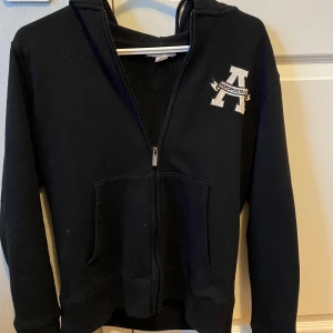 Svart hoodie med dragkedja från Aeropostale - Svart hoodie med dragkedja från Aeropostale. Stor broderad A-logga på bröstet och två fickor framtill. Mjuk insida och huva. Den är i jätte bra skick och är i storlek M. Skriv om du har några funderingar.