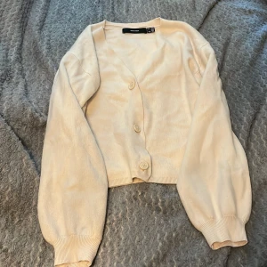Beige kofta från Vero Moda - Säljer en ljusbeige kofta från Vero Moda med tre knappar framtill. Koftan har långa ärmar med breda muddar och är tillverkad i ett mjukt material. Perfekt att slänga på sig när det är lite kyligt ute.