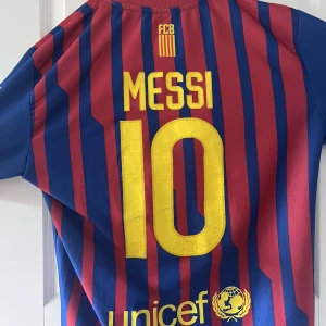 FC Barcelona Messi 10 matchtröja retro - Säljer en klassisk FC Barcelona fotbollströja med Messi 10 på ryggen. Tröjan är blå och röd med gula detaljer, randigt mönster och klubbmärke på bröstet. Perfekt för dig som älskar Barca och Messi!