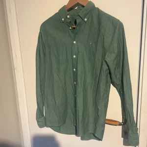 Grön skjorta från Gant - Klassisk grön skjorta från Gant med button-down krage och vita knappar. Liten broderad logga på bröstet. Skjortan har lång ärm och är tillverkad i ett mjukt bomullstyg. Perfekt för en stilren och avslappnad look.