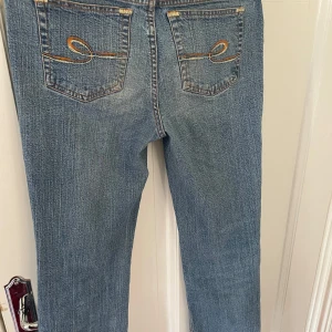 Blå jeans med broderade bakfickor - Säljer ett par klassiska blå jeans med snygga broderade detaljer på bakfickorna och orangea sömmar. Jeansen har normal passform och raka ben, perfekt till vardags. Stängs med dragkedja och knapp framtill. Köpt secondhand men aldrig använt! Vet inte storlek men passar mig som är 165-168 så ungefär 36!!