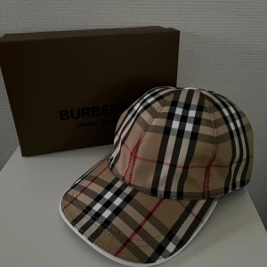  keps från Burberry - Säljer en klassisk rutig keps från Burberry i beige, svart, vit och röd. Kepsen har den ikoniska Burberry-rutiga designen och böjd skärm. Perfekt accessoar för en trendig look.