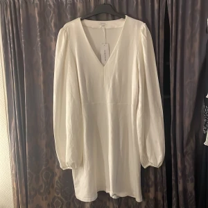 Vit v-ringad klänning från Stradivarius - Säljer en stilren vit klänning från Bubbelroom med v-ringning och långa puffärmar. Klänningen har en enkel, elegant design och är tillverkad i ett mjukt material som ger en fin passform. Perfekt för dig som gillar minimalistisk stil.
