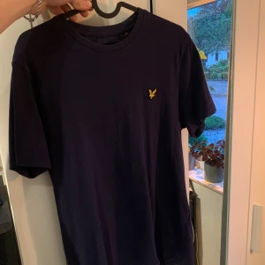 Mörkblå t-shirt från Lyle & Scott - Säljer en mörkblå t-shirt från Lyle & Scott med klassisk rund hals och gul broderad logga på bröstet. T-shirten har normal passform och är tillverkad i mjuk bomull. Perfekt för en stilren och enkel look. Storlek S