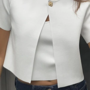 Vit croppad bolerotopp med knapp - Snygg vit croppad bolerotopp med korta ärmar och en dekorativ guldig knapp upptill. Perfekt att styla med jeans för en fräsch look.