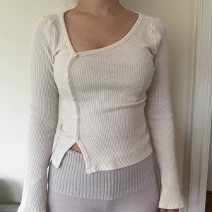 Vit ribbad omlottopp med knappar - Säljer en vit ribbad topp med lång ärm och asymmetrisk omlottknäppning framtill. Tröjan har en unik design med sned knapprad och lätt croppad passform. Perfekt för dig som gillar stilrena och trendiga plagg.