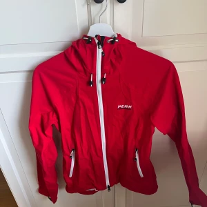 Röd vindjacka från Peak Performance - Röd vindjacka med huva från Peak Performance. Jackan har vita dragkedjor framtill och på fickorna samt justerbar huva. Perfekt för utomhusaktiviteter och har en sportig look.