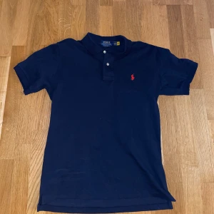 Mörkblå pikétröja från Polo Ralph Lauren - Klassisk mörkblå pikétröja från Polo Ralph Lauren med röd broderad logga på bröstet. Tröjan har korta ärmar, krage och två knappar framtill. Perfekt för en stilren och avslappnad look.