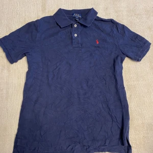 Marinblå pikétröja från Polo Ralph Lauren - Säljer en klassisk marinblå pikétröja från Polo Ralph Lauren med korta ärmar och krage. Tröjan har knappar framtill och den ikoniska röda broderade loggan på bröstet. Perfekt för en stilren och avslappnad look.