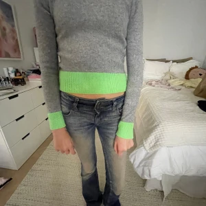 Grå tröja med gröna detaljer - Säljer en grå tröja med ribbade, neongröna muddar vid ärmslut, nederkant och halsringning. Tröjan har en klassisk passform och är långärmad, perfekt för dig som vill sticka ut lite extra.