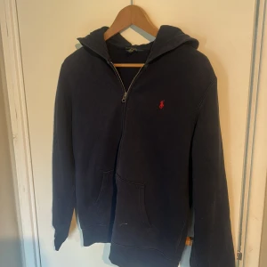 Mörkblå hoodie med dragkedja från Ralph Lauren - Mörkblå ziphoodie från Ralph Lauren med dragkedja framtill och klassisk huva. Tröjan har två fickor och den ikoniska röda loggan broderad på bröstet. Perfekt för en avslappnad stil.