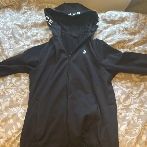 Svart hoodiejacka från Peak Performance - Säljer en svart hoodiejacka från Peak Performance med dragkedja och huva. Jackan har logga på bröstet och text på huvan. Perfekt för träning eller chill. Två fickor med dragkedja framtill.