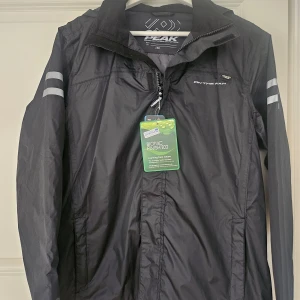 Svart vindjacka från On The Peak - Svart vindjacka från On The Peak med dragkedja och reflexdetaljer på ärmarna. Jackan har hög krage och huva, samt logga på bröstet. Perfekt för utomhusaktiviteter och enkel att bära med sig.