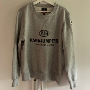 Grå sweatshirt från Parajumpers - Säljer en grå sweatshirt från Parajumpers med svart tryck på bröstet och broderad patch på ärmen. Tröjan har rund halsringning, långa ärmar och ribbade muddar. Perfekt för en avslappnad och stilren look. Pris 350, hör av er vid minsta fundering 