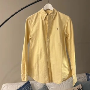 Gul skjorta från Polo Ralph Lauren - Säljer en klassisk ljusgul skjorta från Polo Ralph Lauren i storlek M. Skjortan har button-down krage, broderad logga på bröstet och är tillverkad i bomull. Perfekt för en stilren och tidlös look.