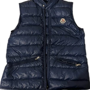 Mörkblå dunväst från Moncler - Snygg mörkblå dunväst från Moncler med quiltad design och dragkedja framtill. Västen har två snedställda fickor med dragkedja och Moncler-logga på bröstet. Perfekt för lager-på-lager och stilren look. Skicket är 10/10 och nypris ligger på ungefär 8000. Ställ gärna frågor. 