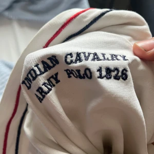 Vit track jacket från Polo Ralph Lauren - Vit track jacket från Polo Ralph Lauren med broderad logga på bröstet och texten 'Indian Cavalry Army Polo 1826' på ryggen. Jackan har röda och marinblå ränder längs dragkedjan och ärmarna samt siffran 3 på ena ärmen.