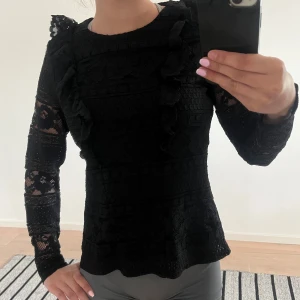 Svart spetsblus med volangdetaljer - Säljer en svart blus med lång ärm i spets och vackra volangdetaljer framtill, baktill och på axlarna. Blusen har ett elegant mönster och är genombruten i materialet, vilket ger en lyxig känsla. Perfekt att styla till både jeans och kjol.