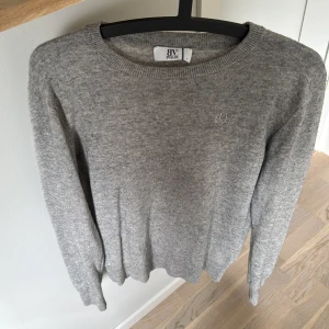 Grå stickad tröja från bvaldi  - Säljer en klassisk grå stickad tröja från bvaldi med rund halsringning och diskret broderad logga på bröstet. Tröjan har långa ärmar och ribbade muddar vid ärmslut och nederkant. Perfekt basplagg till garderoben.