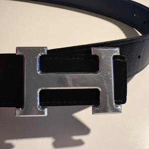 Svart skärp med silverfärgat H-spänne  - Säljer ett stilrent svart bälte med ett stort, silverfärgat H-spänne. Bältet är gjort i skinn och har en klassisk design som passar till många olika outfits. Perfekt accessoar för dig som gillar exklusiva detaljer. Kan vara svart och blått. 