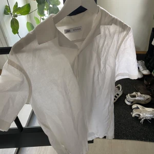 Vit linneskjorta från Zara - Säljer en stilren vit skjorta från Zara i luftig linnemix. Skjortan har korta ärmar, klassisk krage och knäppning framtill. Perfekt för varma dagar när du vill hålla det enkelt men snyggt.