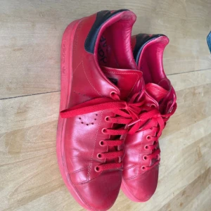 Adidas Raf Simons Stan Smith röda sneakers - Säljer ett par röda Adidas Raf Simons Stan Smith sneakers med svart detalj vid hälen. Skorna är i skinn och har klassisk snörning samt präglad Raf Simons-logga på sidan. Perfekta för dig som gillar statement-sneakers.