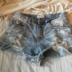 Ljusblå slitna jeansshorts från Acid Blues - Ljusblå jeansshorts med råa kanter och flera slitningar framtill. Klassisk femficksmodell med bälteshällor och knappgylf. Perfekta för en avslappnad stil.