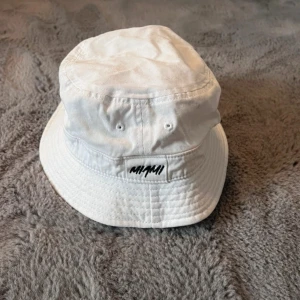 Vit bucket hat med MIAMI-tryck - Säljer en stilren vit bucket hat med svart MIAMI-tryck framtill. Hel men nån fläck jag ej försökt få bort.