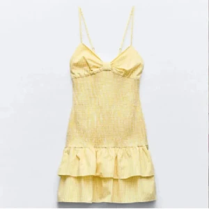 Gul volangklänning med smala axelband - Söt gul kortklänning med smala axelband och volang nertill. Klänningen har en lätt v-ringning och knytning i ryggen. Perfekt för varma dagar och har ett diskret randigt mönster.