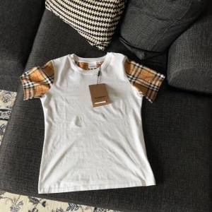 Vit t-shirt med rutiga ärmar från Burberry - Säljer en vit t-shirt från Burberry med klassiska beige-rutiga ärmar. T-shirten har rund halsringning och är kortärmad. Storlek M passar även S 