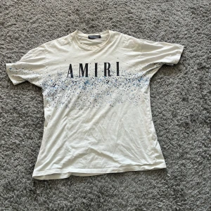 Vit t-shirt från Amiri  - Säljer en vit t-shirt från Amiri med svart logotyp framtill och blått stänkmönster både fram och bak. T-shirten har rund hals och normal passform. 