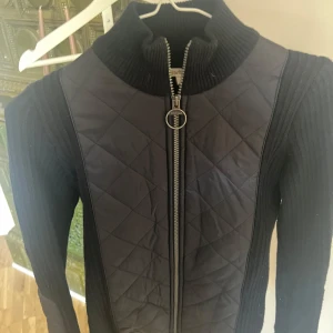 Mörkblå quiltad jacka från Barbour - Säljer en jacka från Barbour med quiltade partier framtill och på ärmarna. Jackan har ribbad krage och ärmar samt dragkedja med rund detalj. 