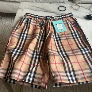 Beige rutiga shorts med snörning - Shorts i beige med klassiskt rutigt mönster i svart, vitt och rött. De har elastisk midja med snörning och sidofickor. Perfekta för sommaren och tillverkade i ett lätt syntetmaterial.