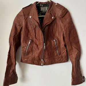 Brun skinnjacka Jofama Marie Serneholt Strl.34  - Snygg brun skinnjacka från Jofama by Marie Serneholt. Jackan har coola detaljer som silvriga dragkedjor, axelpartier med struktur och knappar. Fodret har ett unikt blommigt mönster i guld och svart. Perfekt för dig som vill ha en edgy look. Ficka med dragkedja på insida. Nyskick! Nypris: 2795kr 