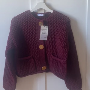 Vinröd stickad kofta från Zara - Säljer en chunky vinröd stickad kofta från Zara med stora guldiga knappar och två fickor framtill. Koftan har lång ärm och en loose passform, perfekt för lager på lager. Superfin till höst och vinter!