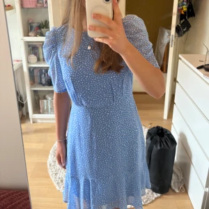Blå prickig klänning från Vero Moda - Supersöt blå kortklänning från Vero Moda med vita prickar, puffiga kortärmar och markerad midja. Klänningen har volang nertill och dragkedja på ryggen. Perfekt för sommarens alla tillfällen!