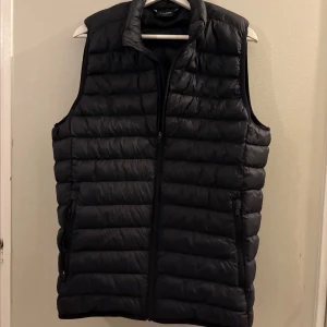 Svart dunväst med dragkedja - Säljer en svart dunväst från Everest med quiltad design och hög krage. Västen har två fickor med dragkedja och stängs med en hel dragkedja framtill. Perfekt att slänga över en hoodie eller tröja för extra värme.