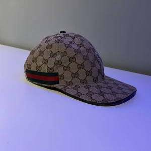 Gucci keps med GG-mönster och röd/grön rand - Snygg keps från Gucci i beige och brunt GG-monogram över hela kepsen. Klassisk böjd skärm och justerbart spänne bak. Sidan har den ikoniska röd-gröna randen. Tillverkad i bomullscanvas med svart insida. Perfekt för dig som vill sticka ut med lyxig streetstyle.