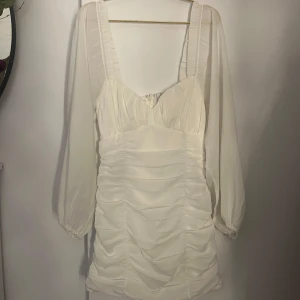 Vit kortklänning från Nelly - Supersöt vit kortklänning från Nelly med puffiga långärmar och snygg rynkad effekt över hela klänningen. Djup fyrkantig ringning och dragkedja bak. Använt 1 gång till skolavslutningen 