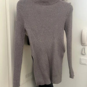 Ljusgrå ribbad polotröja med slits - Säljer en ljusgrå ribbstickad polotröja med lång ärm och hög krage. Tröjan har snygga slitsar i sidorna och en figurnära passform. Perfekt till lager-på-lager och enkel att styla med jeans eller kjol. Den är lite söndrig på ena sidan vilket funkar om man bara syr igen den. 