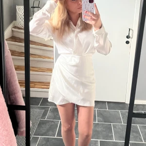 Vit omlottklänning i satin - Snygg vit kort omlottklänning med skjortkrage och långa ärmar. Klänningen har en lätt glansig satinlook och markerad midja som ger en fräsch och stilren vibe. Perfekt för dig som gillar en clean och chic stil.