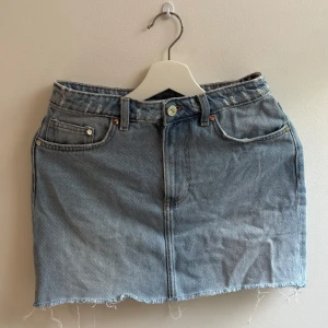 Ljusblå jeanskjol med rå kant - Säljer en ljusblå jeanskjol, med klassisk femficksdesign och rå, fransig kant nertill. Kjolen har bälteshällor, knapp och dragkedja framtill samt bakfickor. Perfekt för dig som gillar en avslappnad och trendig stil.