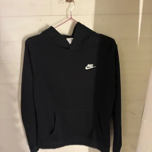 Svart Nike hoodie XL - Svart hoodie från Nike i storlek XL barn med klassisk vit logga på bröstet. Tröjan har huva, lång ärm och en stor magficka framtill. Perfekt för dig som gillar en enkel och stilren look. Materialet är mjukt och skönt