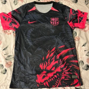 FC Barcelona x Nike svart draktröja - Unik FC Barcelona fotbollströja från Nike med svart bas och mörkgrått drakmönster över hela tröjan. Rosa detaljer på ärmar, axlar, Nike-logga och klubbmärke. Tillverkad i lätt och ventilerande polyester, perfekt för dig som vill sticka ut på planen.