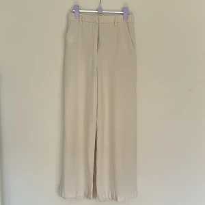 Beiga vida viskosbyxor  - Snygga beiga byxor i tunnt material med vida ben och hög midja. Byxorna har bälteshällor och sidofickor, perfekta för en chill och stilren look. Lätta och luftiga, passar grymt till varma dagar.